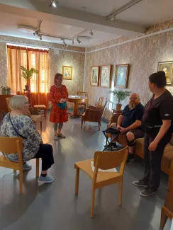 vanha 1920 luvun tyyliin sisustettu huone jossa tauluja seinällä ja vanhoja huonekaluja hymyilevä henkilö puhuu keskellä huonetta ihmisille.