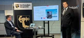 Skeleton Technologiesin edustustoa, vasemmalla mies istuu tuolilla, oikealla mies puhuu ja katsoo kameraan. Taustalla näkyy Varkauden kaupungin ja Skeleton Technologiesin roll-upit sekä esitys.