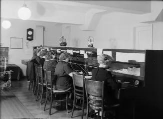 Puhelinkeskus jossa monta työntekijää puhelinvälityspöytien ääressä välittämässä puheluita